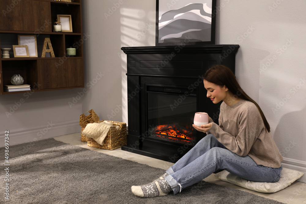 Poster beautiful young woman with cup of hot drink sitting on floor near fireplace at home. space for text