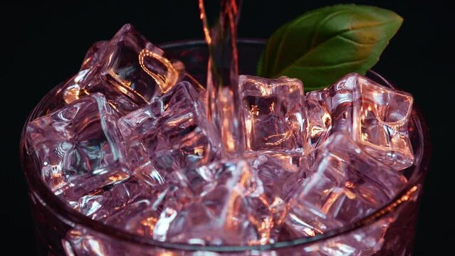 Static shot of artistic cocktail with light red or pink light and coloring being poured over ice with a black background and a green leaf garnish