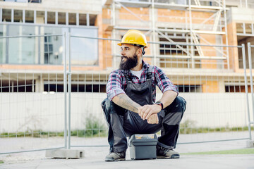 A worker sitting on a toolbox and waiting for a ride at construction site.