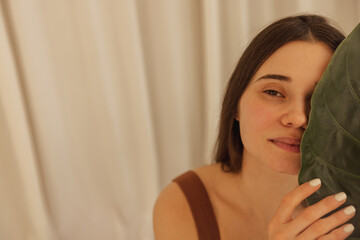Naklejka premium Close up beautiful caucasian young woman with natural makeup and clean fresh healthy skin covers her face with palm leaf. Brunette looks at camera, posing against background of white wall. Spa concept