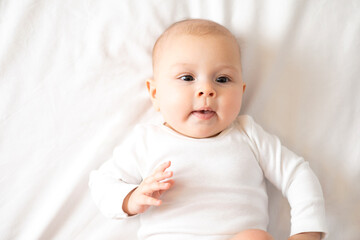 Portrait of a cute baby girl of 5 months in a white bodysuit on a white bed in the bedroom. Space for text. Child lifestyle