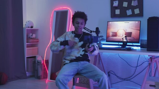 Slowmo of 14 year old Biracial online streamer recording video of himself talking to subscribers on smartphone while playing first person shooter game on computer in his room