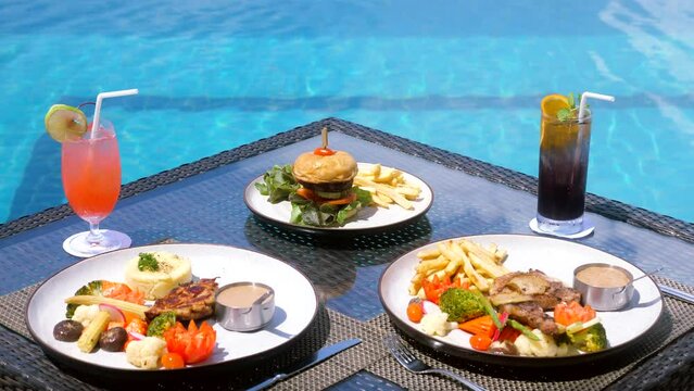 Dinner with cold cocktails near hotel swimming pool. Exotic summer party. Tropical beach lifestyle, vacation. Table with plates of healthy food, meat and vegetables poolside