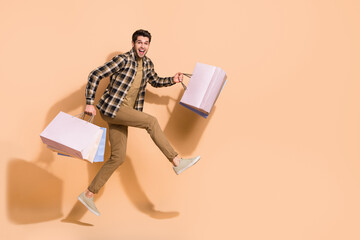 Full length body size view of attractive cheerful crazy guy jumping carrying bags isolated over beige pastel color background