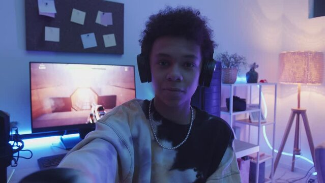 Handheld POV slowmo of Biracial teenage gamer in headphones talking at camera while recording video blog of himself playing fps game on pc at home