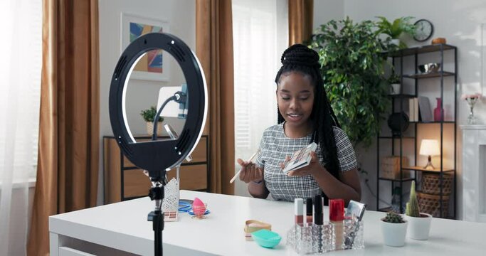 Live streaming and blogging concept. Smiling vlogger does makeup, showing eye shadow palette, smokey eyes tutorial, influencer looks at cell phone camera on tripod with ring light.
