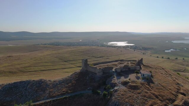 Long drone tracking shot of Enisala, medieval fortress on top of a hill surrounded by lakes and plains on a clear sunny day