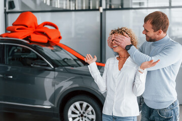 Closing her eyes by hands. Husband makes gift for his wife. Modern new car