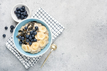 Oatmeal porridge bowl with banana slices, blueberries and pepita seeds. Grey cement texture background. Top view, copy space for text or design elements