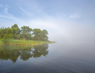 Mystical landscape. Fog in the early morning on the river. The trees near the water are illuminated by the rays of the rising sun.