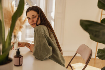 Beautiful young caucasian girl sits at table with care cosmetics, looks at camera. Brunette model in shirt holds jar of product. Beauty, skin, lifestyle concept
