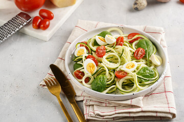 Keto paleo low-carb zoodles: zucchini noodles with parmesan cheese, cherry tomatoes, quail eggs on a checkered kitchen towel light grey surface