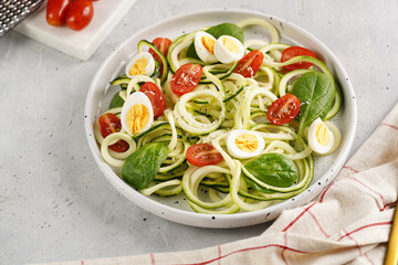 Keto paleo low-carb zoodles: zucchini noodles with parmesan cheese, cherry tomatoes, quail eggs on a checkered kitchen towel light grey surface