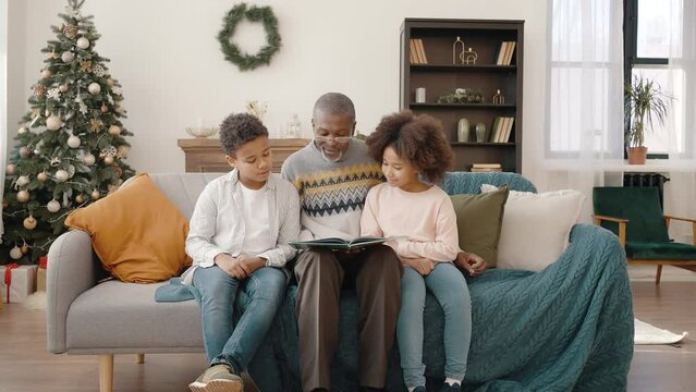 Senior african american grandfather reading fairy tale to grandchildren, sitting together at home with Christmas tree