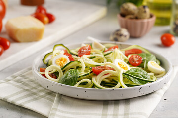 Keto paleo low-carb zoodles: zucchini noodles with parmesan cheese, cherry tomatoes, quail eggs on a checkered kitchen towel light grey surface