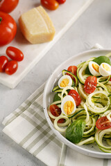 Keto paleo low-carb zoodles: zucchini noodles with parmesan cheese, cherry tomatoes, quail eggs on a checkered kitchen towel light grey surface