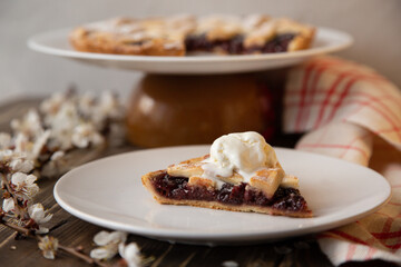 American cherry pie. A piece of  pie with a scoop of ice cream on a white plate in the foreground with a sprig of cherry blossoms. 