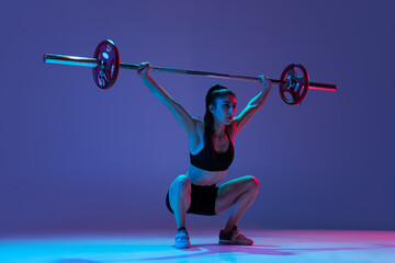 Naklejka na ściany i meble Portrait of muscled woman in sportswear training with a barbell isolated on purple background in neon light. Sport, weightlifting concept