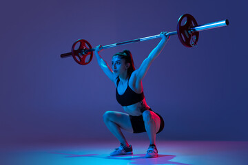 Portrait of muscled woman in sportswear training with a barbell isolated on purple background in neon light. Sport, weightlifting concept