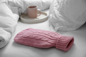 Hot water bottle with knitted cover and cup of tea on bed