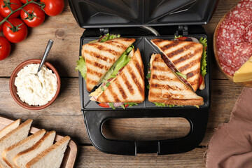 Modern grill maker with sandwiches and different products on wooden table, flat lay