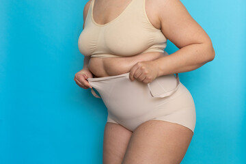 Plus size woman in beige underclothes pulling shapewear panties over belly closeup. Tighten figure...