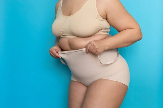 Plus size woman in beige underclothes pulling shapewear panties over belly  closeup. Tighten figure and wear lifting underwear for weight loss, blue  background. Concept of diet and fighting overweight. Stock Photo | Adobe  Stock