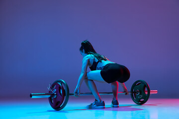 Portrait of muscled woman in sportswear training with a barbell isolated on purple background in...