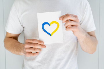 partial view of man in white t-shirt holding card with blue and yellow heart on grey background.