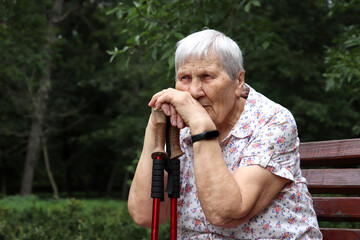 Portrait of elderly gray-haired woman sitting with walking sticks on a bench in a park. Healthy lifestyle in old age, life in retirement
