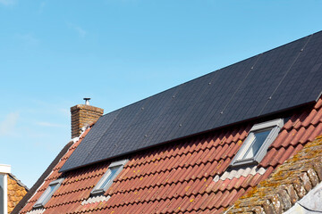 A row of dark solar panels on a house roof with orange tiles against a blue sky.     