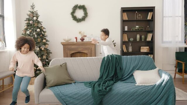 Happy african american boy and girl running around sofa together at decorated living room, playing catch-up at home