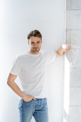 man in white t-shirt standing with hand in pocket of jeans near white wall.