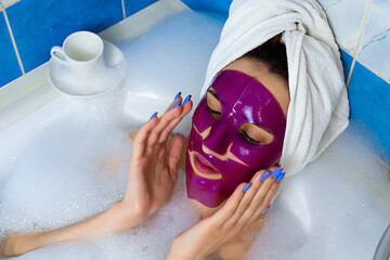Caucasian young red-haired woman relaxing in bathroom, wearing bath towel wrapped around head, lying in a foam bathtub with pink clay face mask on face, enjoying her nail manicure. Body and Skin Care