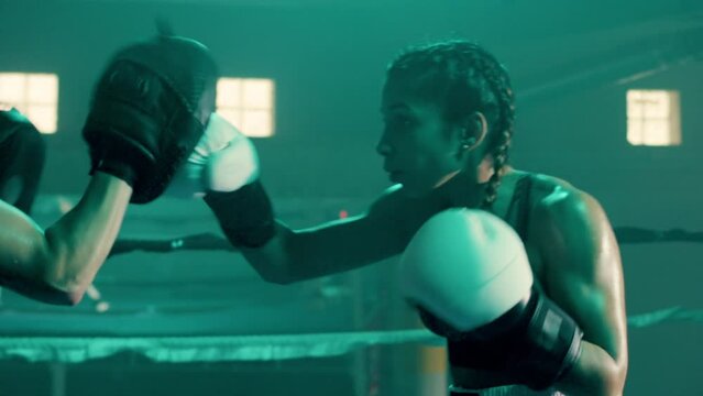 Medium shot of strong young woman training with coach, wearing boxing gloves and punching boxing paws. Attractive female boxer training indoors in gym. Sport, boxing concept