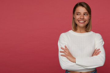 Studio portrait of young female student posing over red background, smiling broadly, looks aside at copy space, keeps her hands folded,