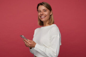 Studio portrait of young female student posing over red background, smiles broadly while typing message on her phone