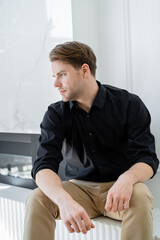 stylish and dreamy man in black shirt sitting at home and looking away.