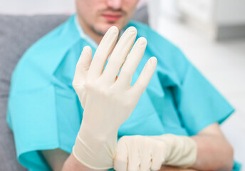 Young caucasian male patient in medical disposable pajamas puts sterile surgical white gloves on...
