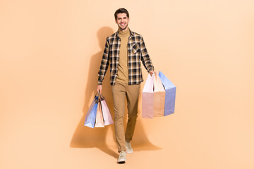 Full length body size view of attractive cheerful guy carrying bags new things isolated over beige...