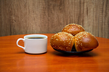 Cup of coffee and buns on a wooden background. White coffee cup on a wooden background. Buns with sesame seeds lie on a plate. A cup of black coffee. Coffee and goodies.