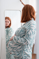 pregnancy redhead woman in pajamas looking at mirror at her belly 