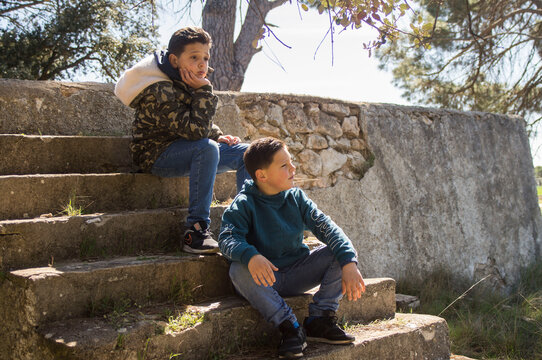 Adventurous kids sitting on valsa steps in a natural environment with blue sweatshirt and camouflage jacket.