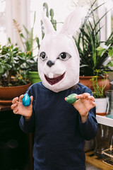 Girl celebrating Easter with a rabbit mask and some chocolate eggs in a glassed-in area surrounded by plants. Easter celebration and costume concept.