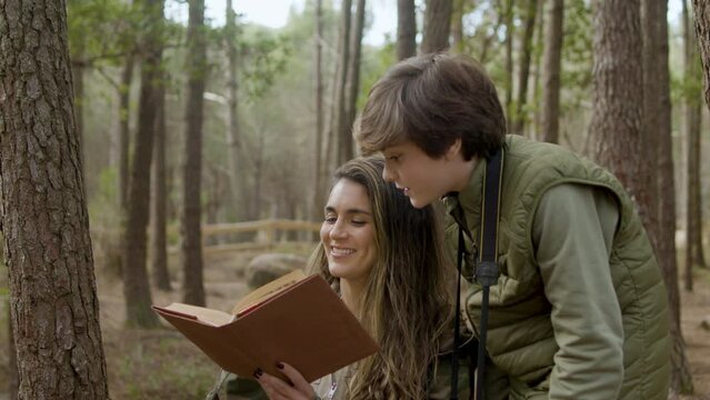 Happy mother and her young son exploring nature in pine forest. Long-haired Caucasian woman holding book while brown-haired boy looking into it and reading out loud. Nature, education concept