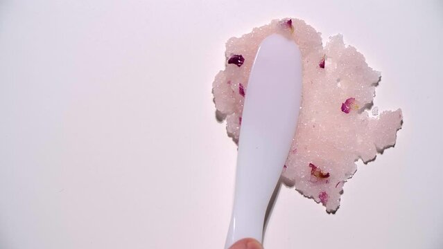 Close-up of the oily texture of a pink body scrub with rose petals and salt crystals is smeared with a white plastic spatula on a smooth matte white background. High quality 4k footage