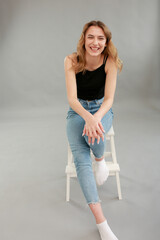 Beautiful girl model appearance in light jeans and black T-shirt on a gray studio background. Girl model sitting on a white chair. Beautiful blonde shows poses for a photo shoot
