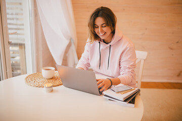 Young woman freelancer working online using her laptop, drinking tea in a cozy wooden house. Work from home.