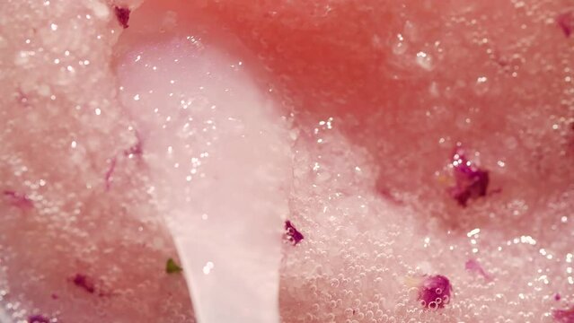 Close-up of the oily texture of a pink body scrub made of sea salt and rose petals slow stirring with a white plastic spatula. High quality 4k footage
