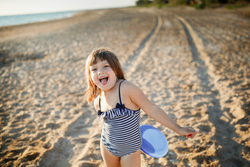 Cute european child girl in a swimsuit runs on the sand and plays frisbee on the beach. Summer holidays and adventures. Active games on the sea coast.